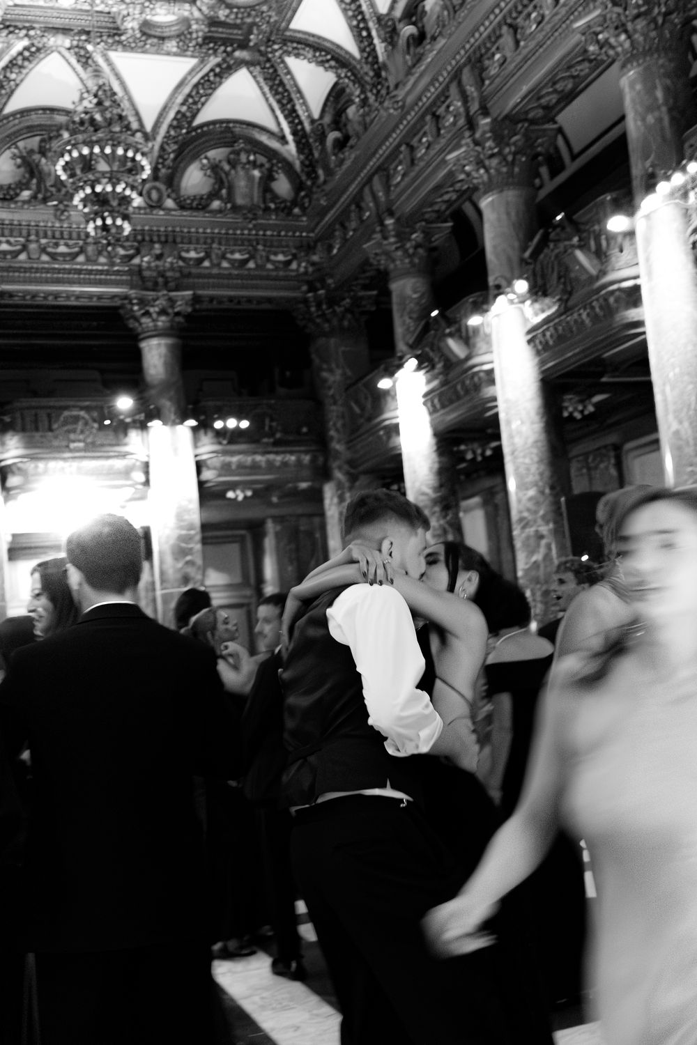 Black and white photo of elegantly dressed guests dancing in an ornate ballroom with grand columns and arched ceilings.
