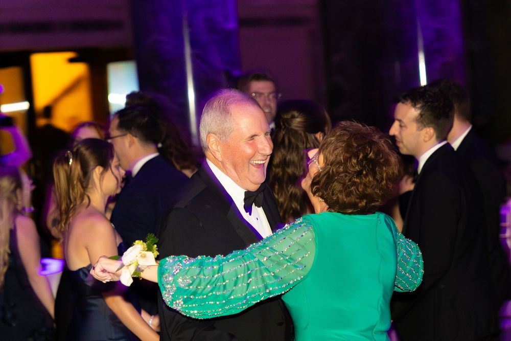 Elderly couple dancing joyfully at a formal gala event, woman in green sequin dress, man in tuxedo with bow tie.