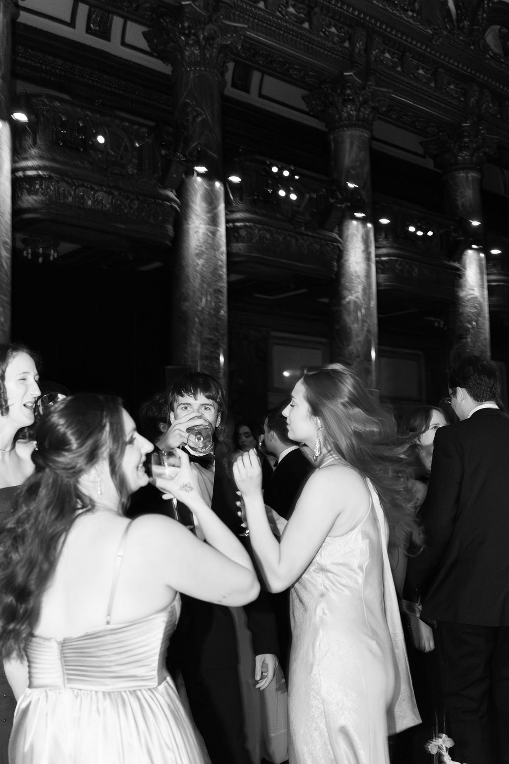 Black and white photo of elegantly dressed guests dancing at a formal masquerade ball in a grand venue.