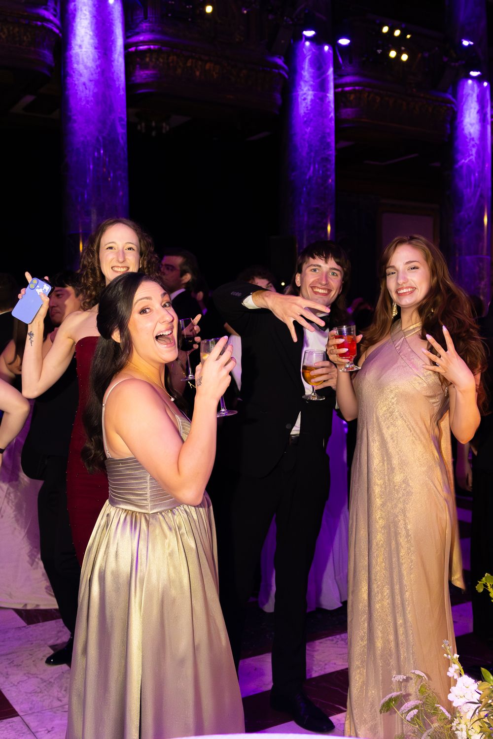 Elegantly dressed young adults celebrate at a formal gala event with purple-lit marble columns in the background.