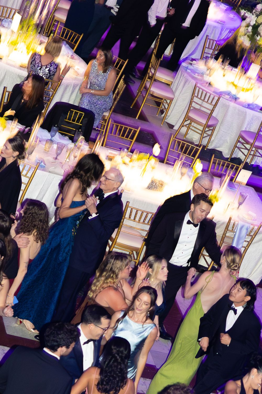 Elegant formal gala event with guests in evening wear dining and socializing at candlelit tables in a grand ballroom.