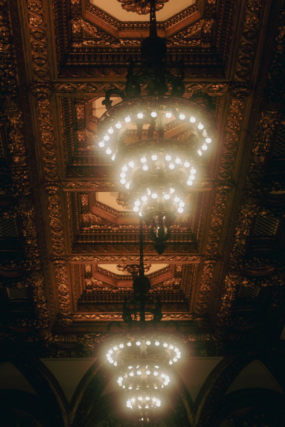 Ornate golden ceiling with elegant chandelier rows glowing warmly in a grand historic venue.