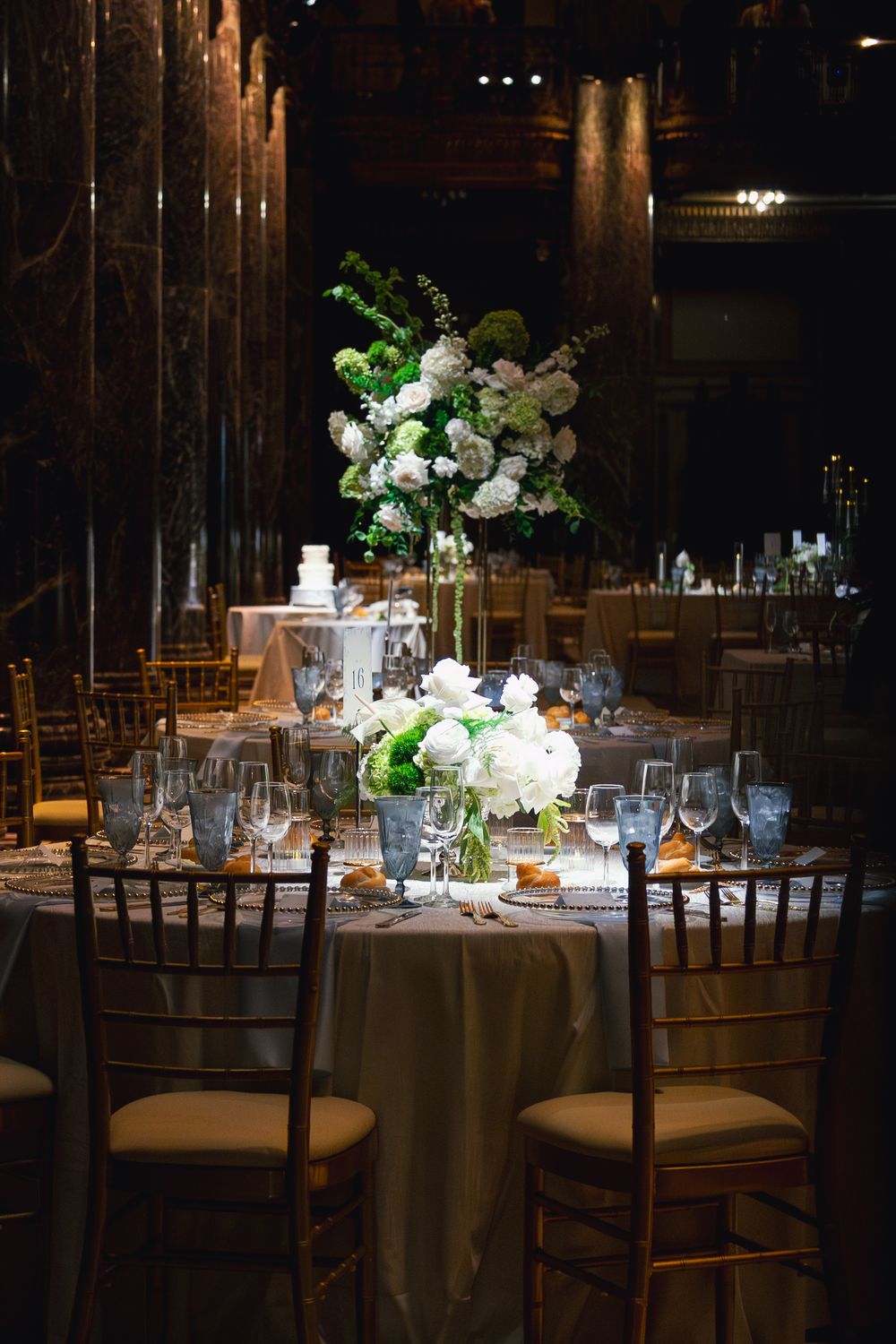 Luxurious wedding reception table with tall white floral centerpiece, gold chiavari chairs, and crystal glassware in dark venue.