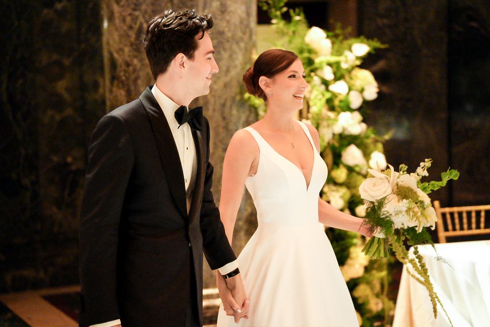 Bride and groom holding hands and smiling at wedding ceremony with floral arrangements in background.