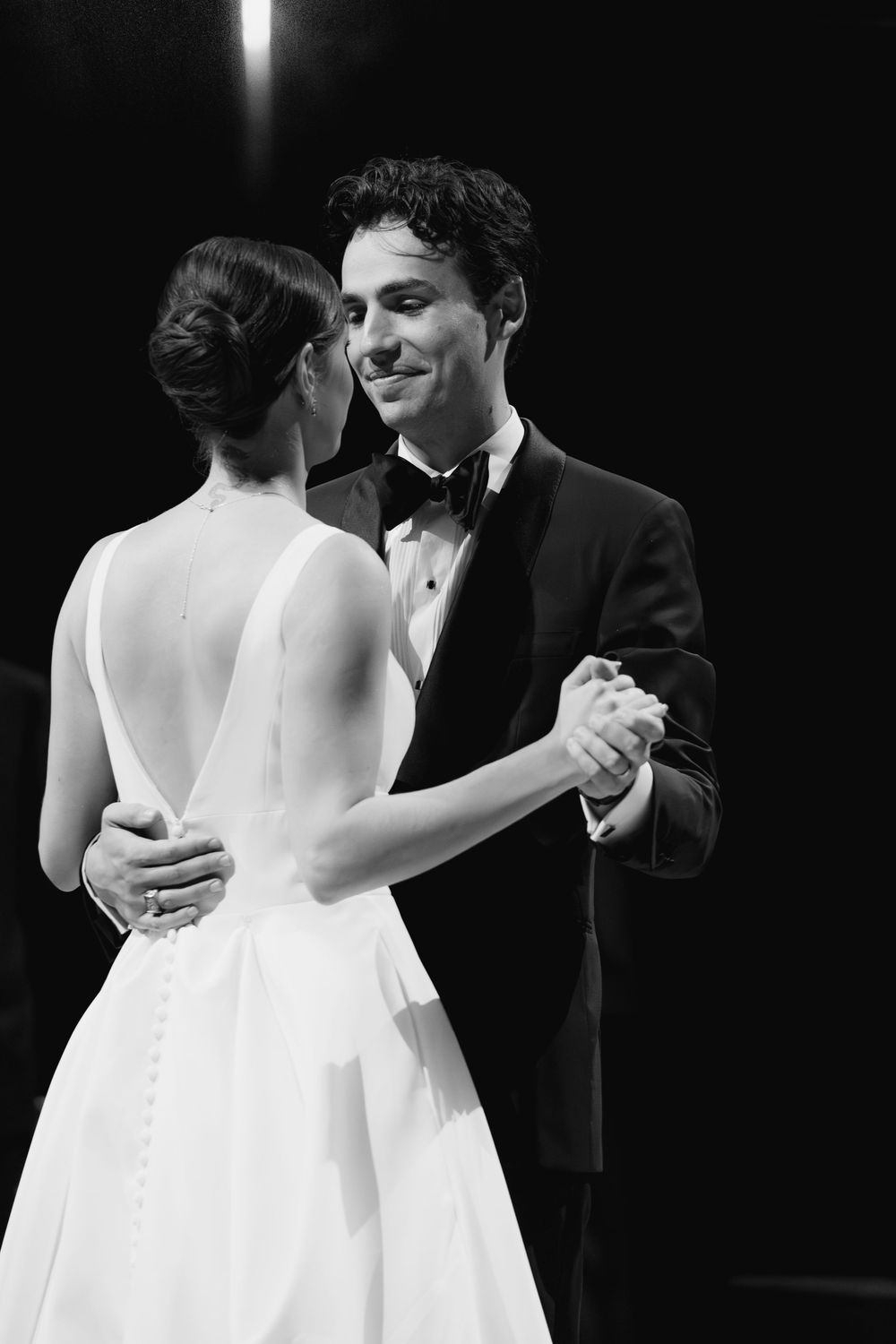 Black and white photo of a smiling groom in a tuxedo sharing a first dance with his bride in a backless white gown.