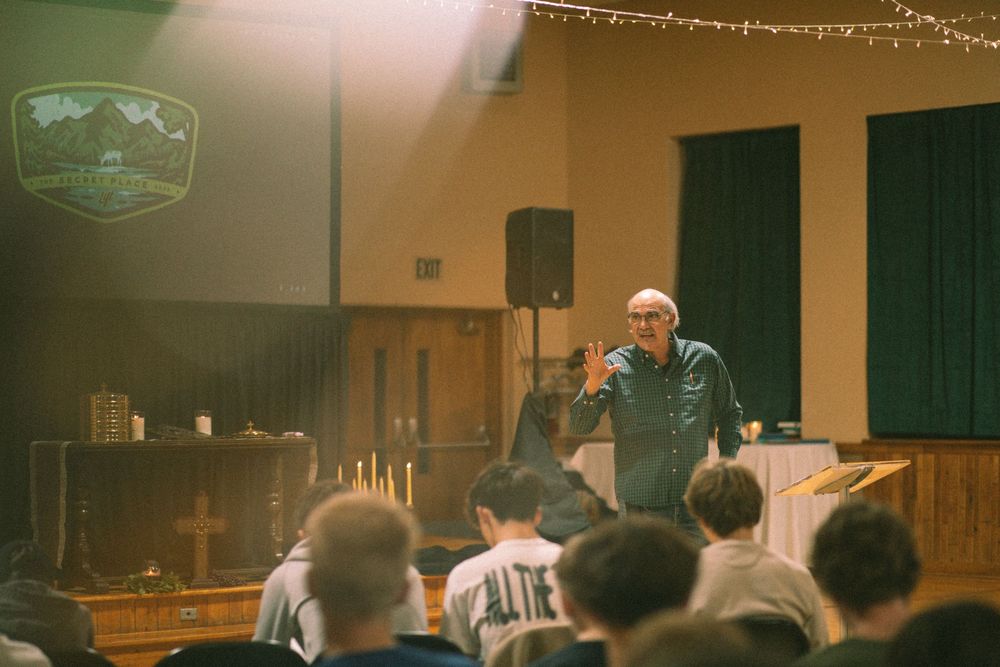 A speaker addresses a seated audience in a warmly lit hall with a projected logo on screen.
