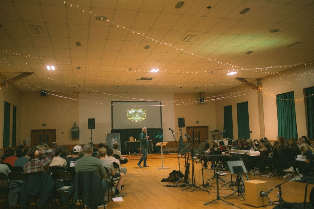 A wide view of an indoor church gathering with a speaker, projection screen, and string lights.