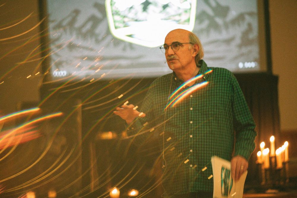 A man in a green checkered shirt speaks at a church event surrounded by candles and warm light streaks.