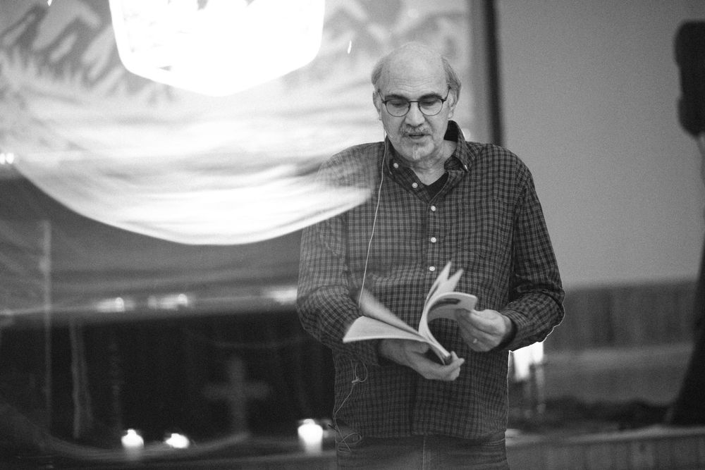 A man with glasses reads from an open book while speaking at a candlelit church event in black and white.