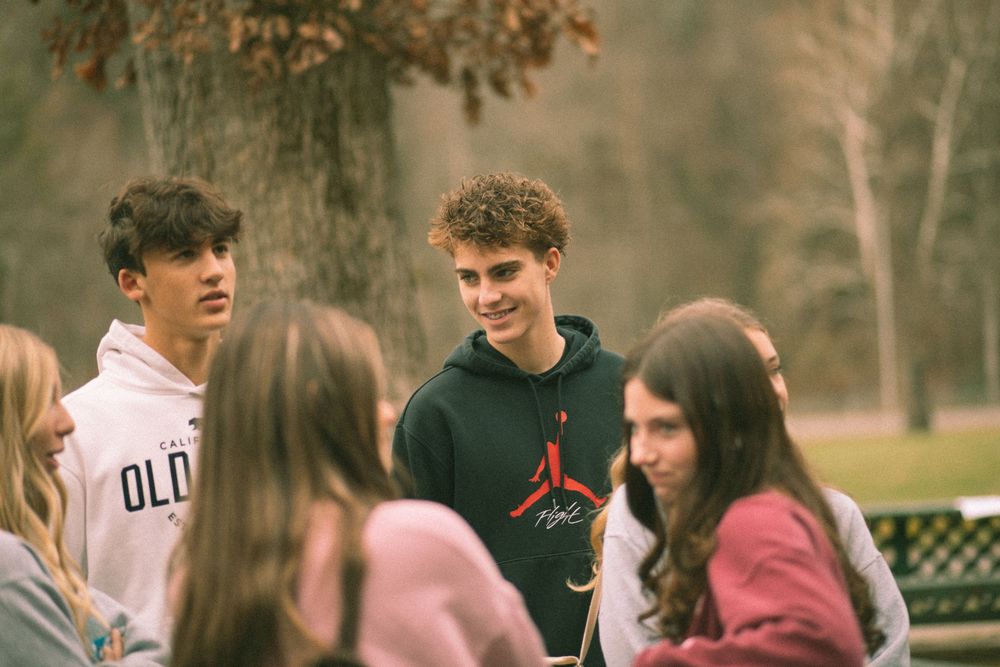Group of teenagers in hoodies socializing outdoors near trees during a youth retreat event.