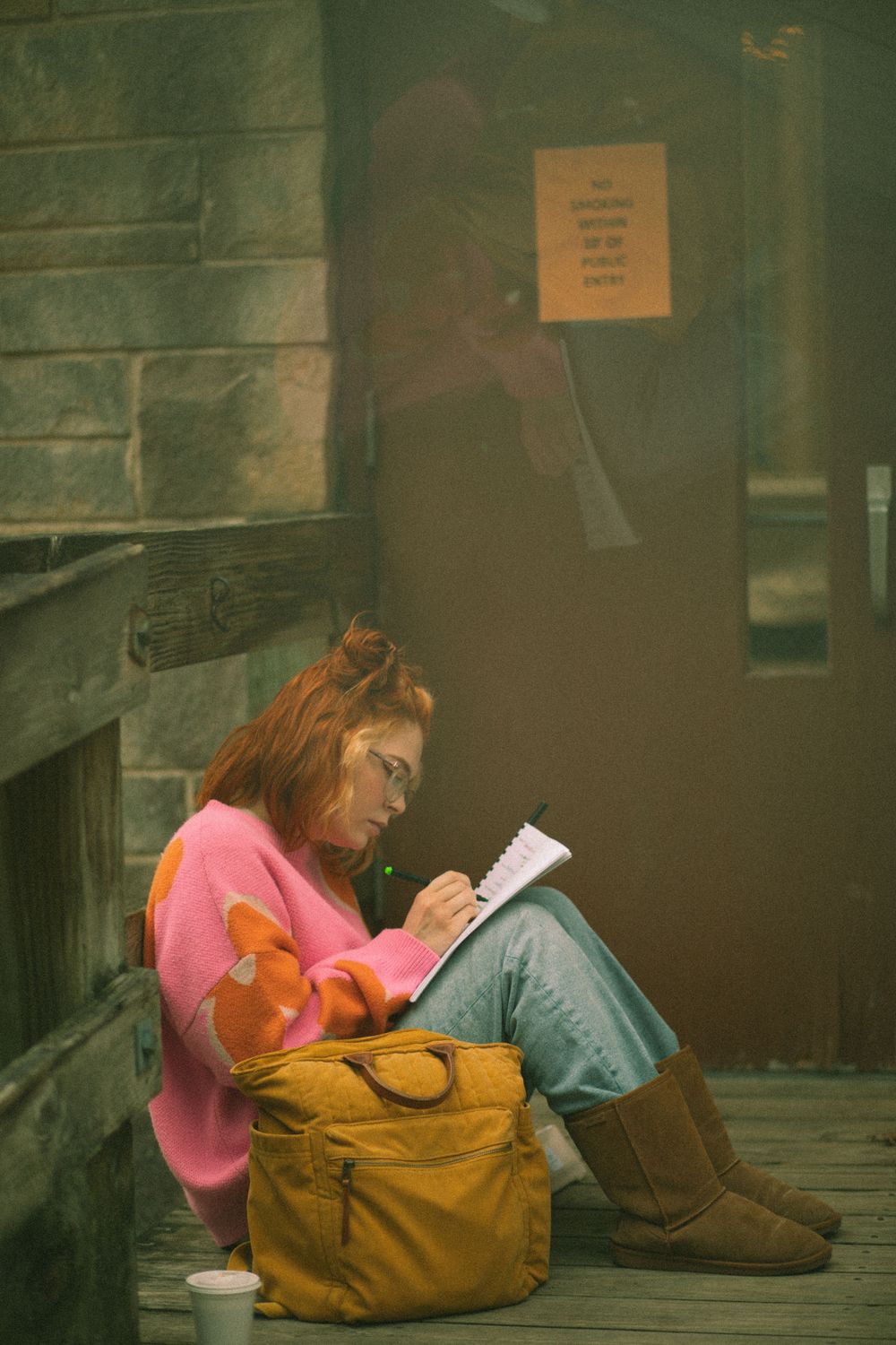 Young woman in pink hoodie sits on wooden steps writing in a notebook with a bag beside her.