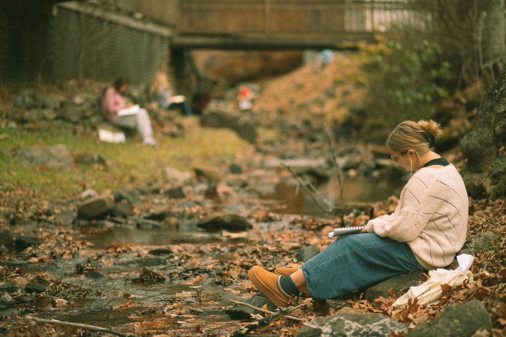 Woman sitting on rocky creek bed reading book surrounded by fall foliage under bridge.