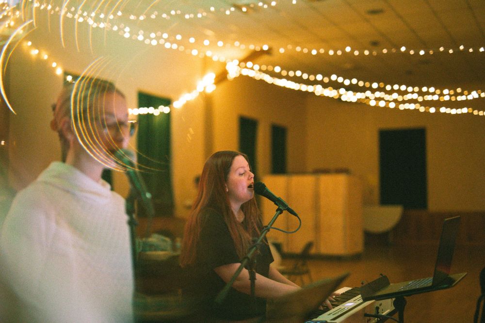 Female vocalist singing passionately at keyboard with string lights glowing in background.