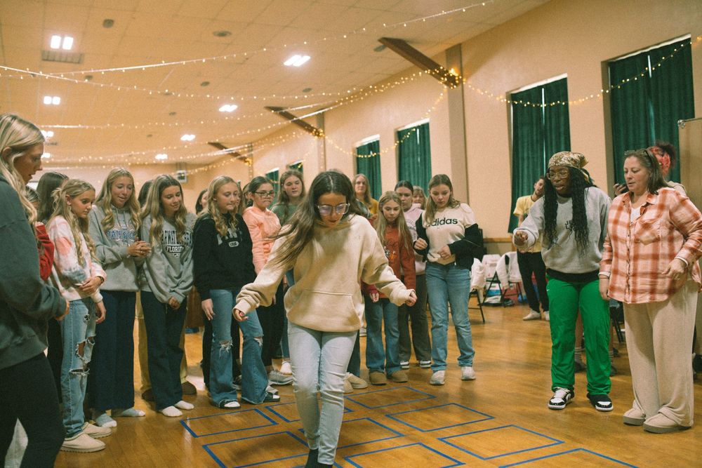 Group of teenage girls participate in a circle dance activity in a decorated church gymnasium.