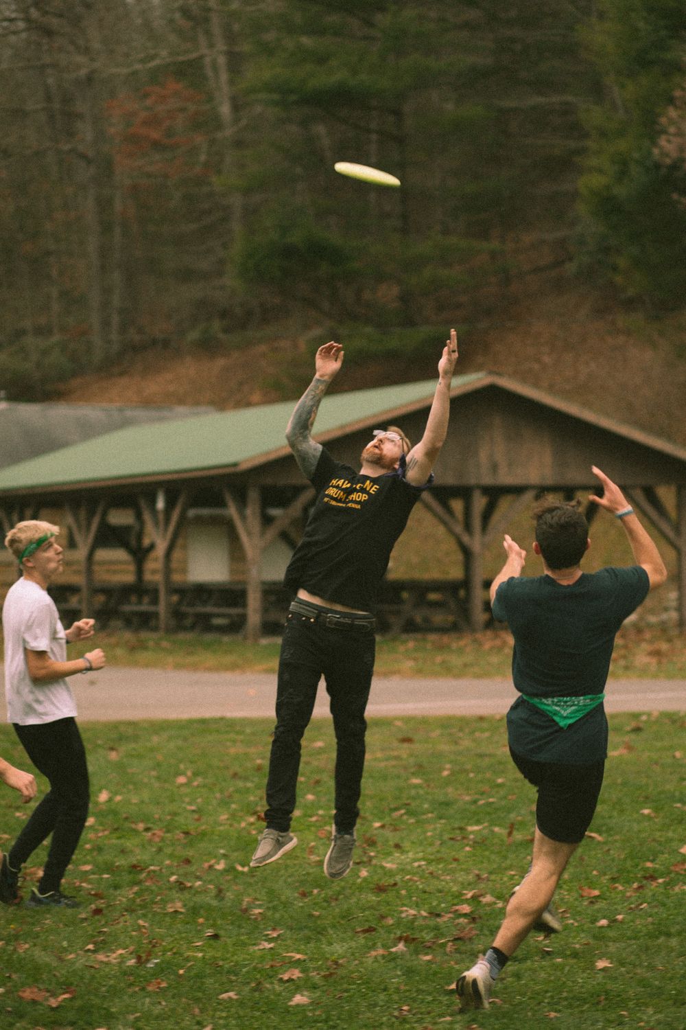Two men jumping to catch a frisbee outdoors near a rustic wooden pavilion.