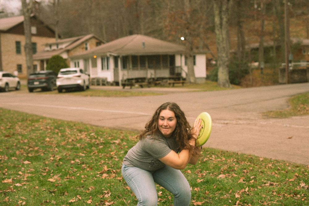 Young woman crouching to catch a yellow frisbee outdoors in a grassy yard with houses in background.