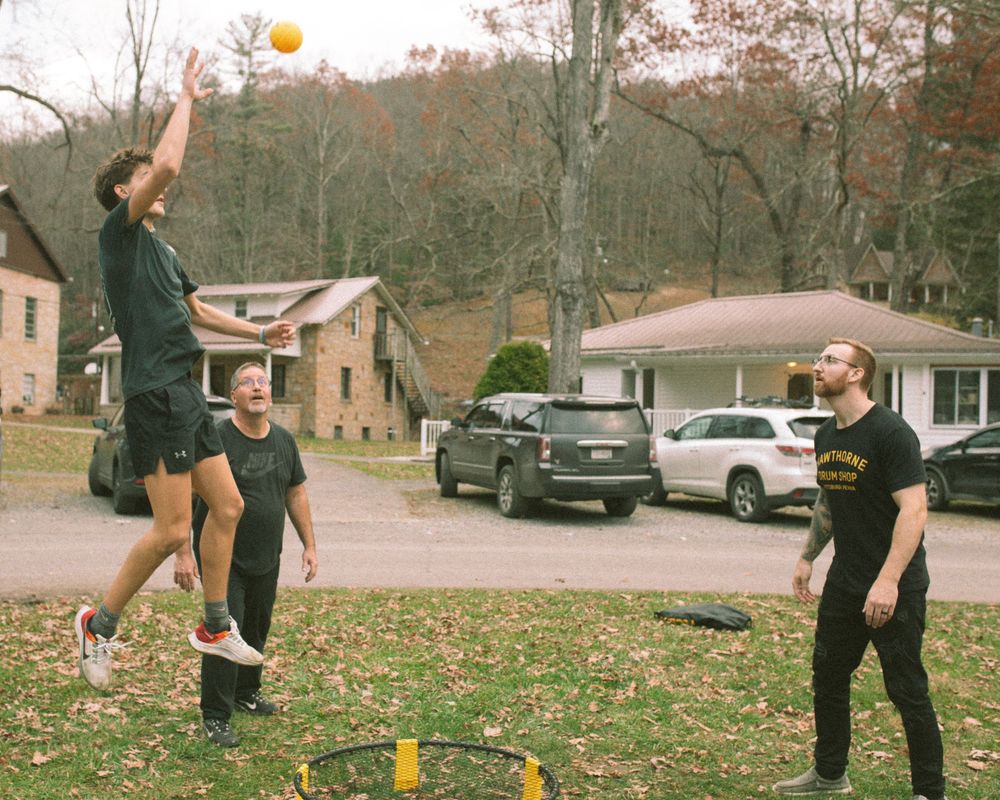 Three men playing spikeball in a residential driveway surrounded by autumn foliage.