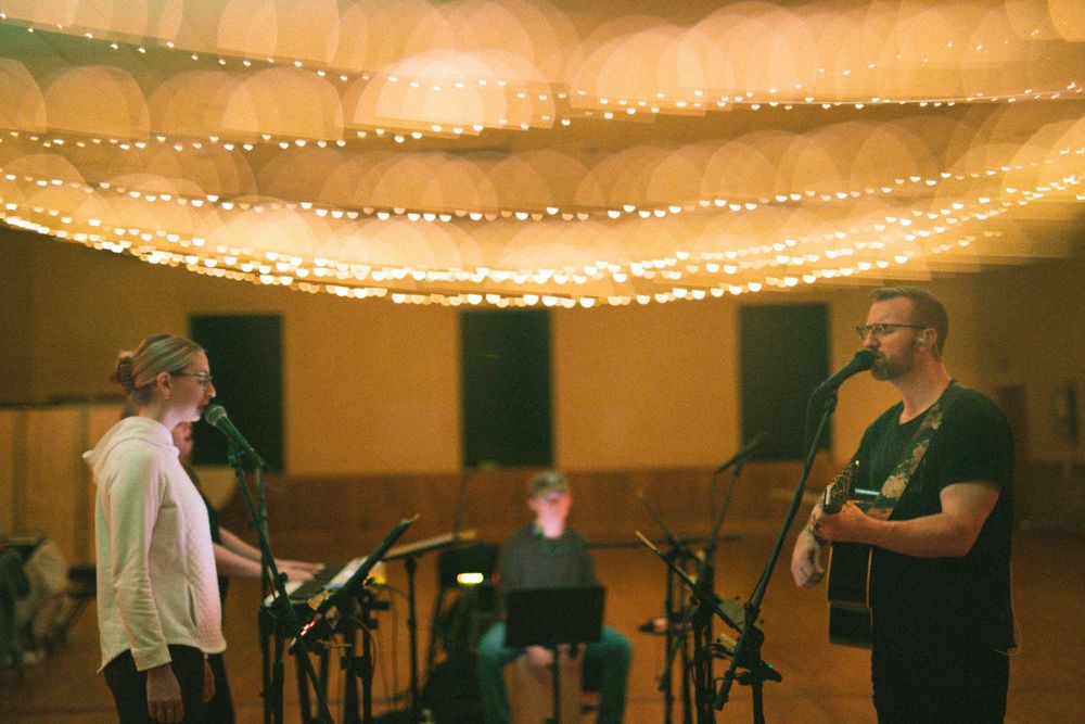 Male guitarist and female vocalist performing together under warm string lights in hall.
