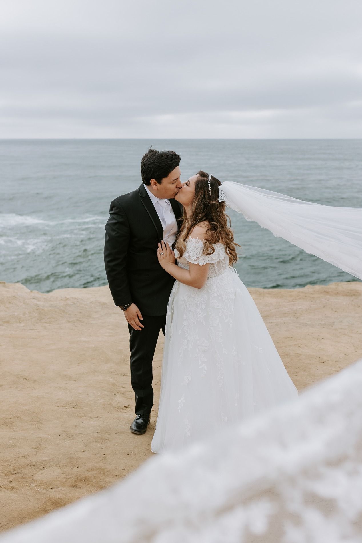 Bride and groom share a kiss on a coastal cliff, her veil flowing in the wind, with the ocean in the background.