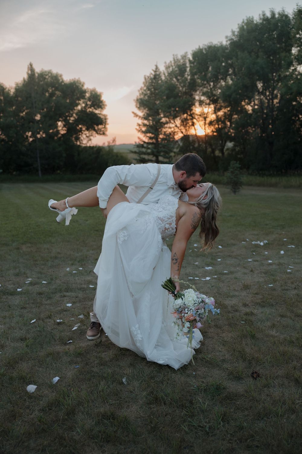 Groom dips bride for a romantic sunset kiss outdoors, bride holding a pastel bouquet in a white gown.