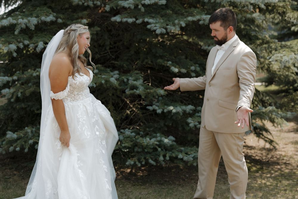 Bride in lace wedding gown and veil shares first look moment with groom in tan suit outdoors near evergreen trees.
