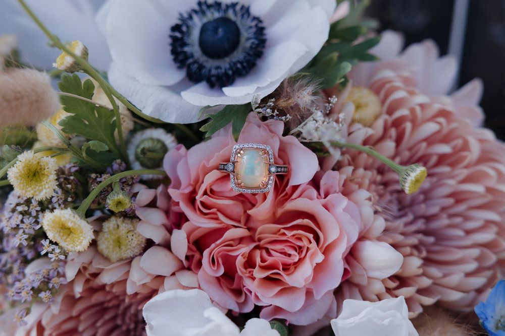 Close-up of elegant opal and sapphire rings resting on a lush bridal bouquet with pink roses and anemones. Bismarck, ND photographer