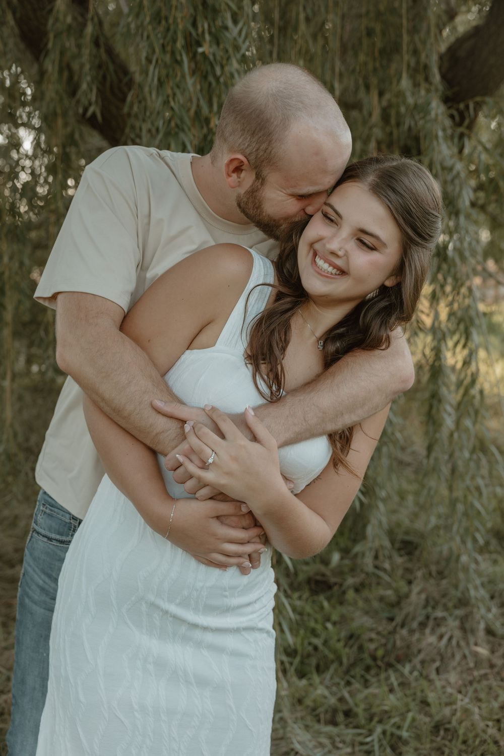 Man embracing woman in white dress from behind, both smiling under a weeping willow tree outdoors. Bismarck, ND photographer