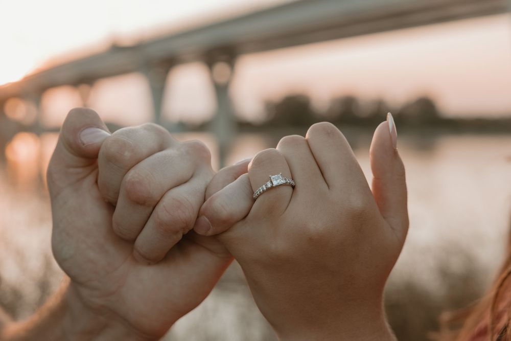 Two hands linking pinky fingers with an engagement ring visible, set against a blurred bridge and sunset backdrop. Bismarck, ND photographer