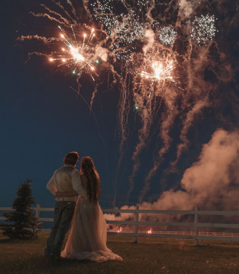 Couple in wedding attire watching colorful fireworks light up the night sky at an outdoor celebration. Bismarck, ND photographer