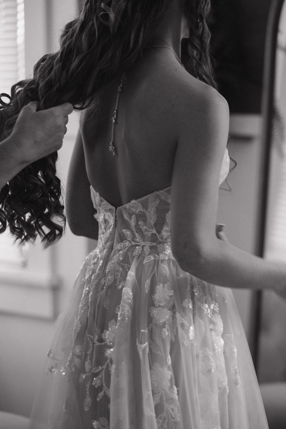 Black and white photo of a bride in a backless floral wedding dress being helped by an assistant. Bismarck, ND photographer