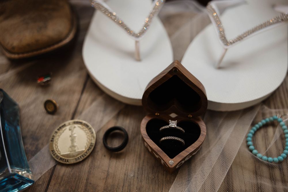 Wedding rings in heart-shaped box beside flip flops, bracelet, and coins on wooden surface. Bismarck, ND photographer