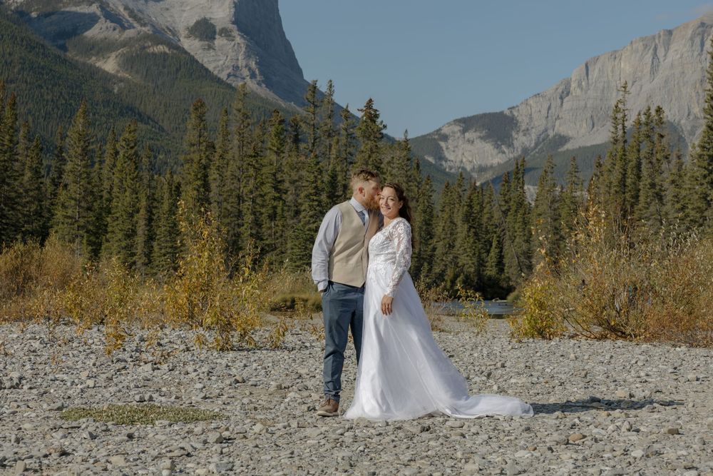 Bride and groom pose on rocky riverbed with stunning mountain backdrop during outdoor fall wedding in Banff. Bismarck, ND travel photograph