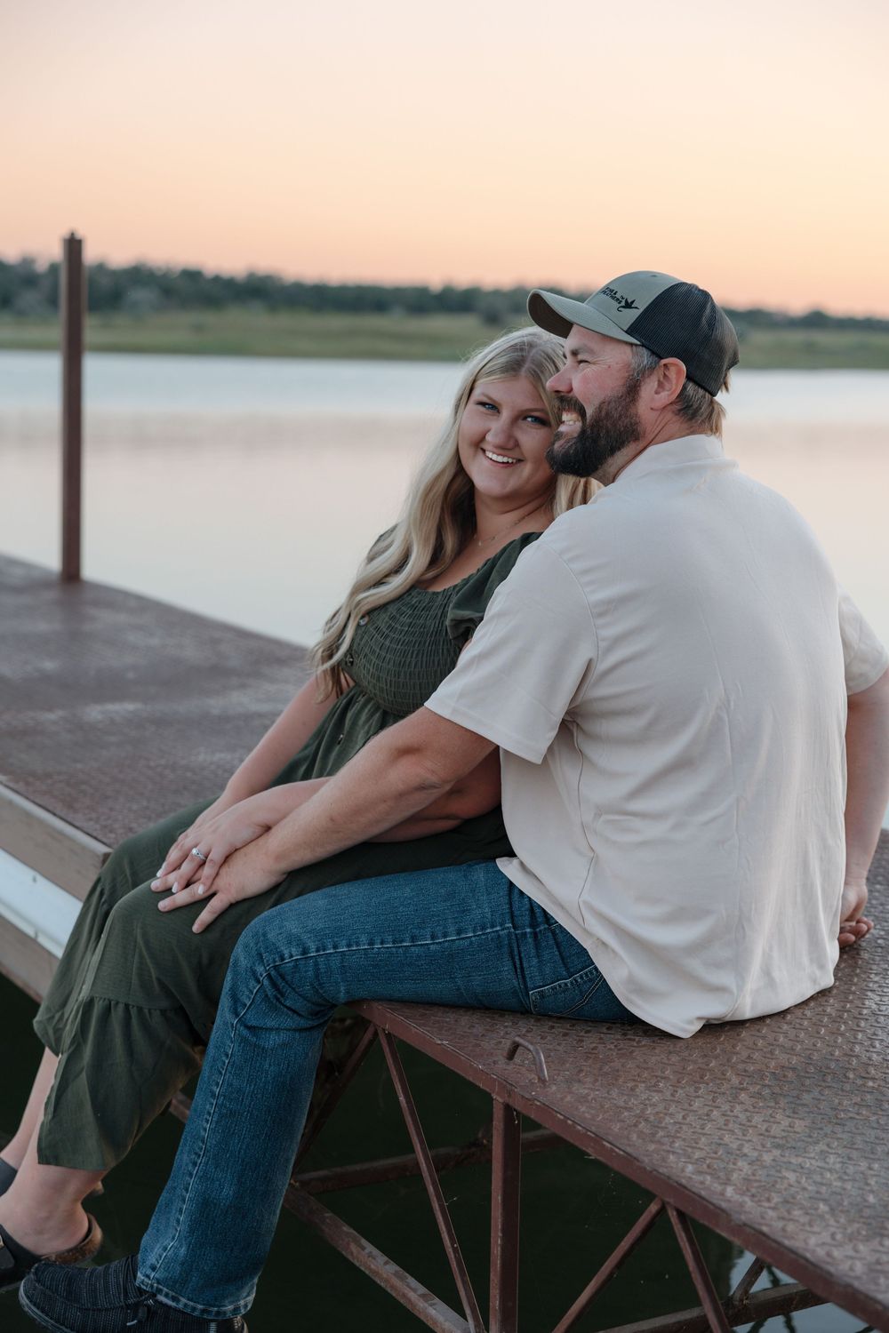 Couple sitting on a dock at sunset, woman in green dress smiling as man in white shirt and cap kisses her cheek. Garrison, ND Photographer