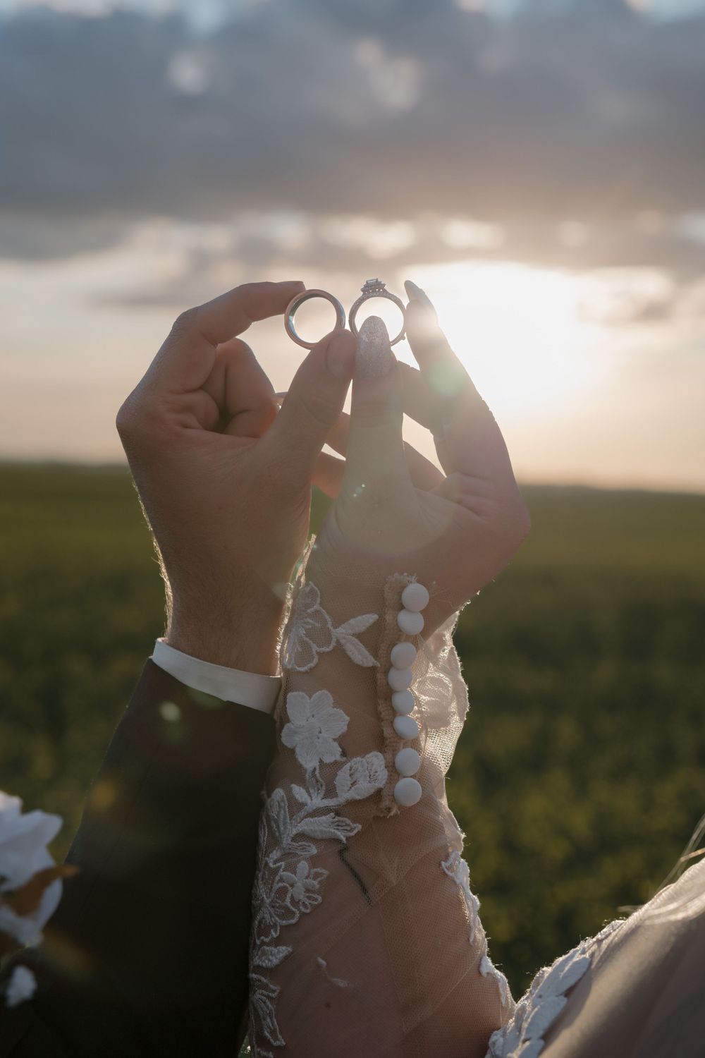 Bride and groom holding wedding rings up toward golden sunset sky above green fields. Garrison, ND Photographer