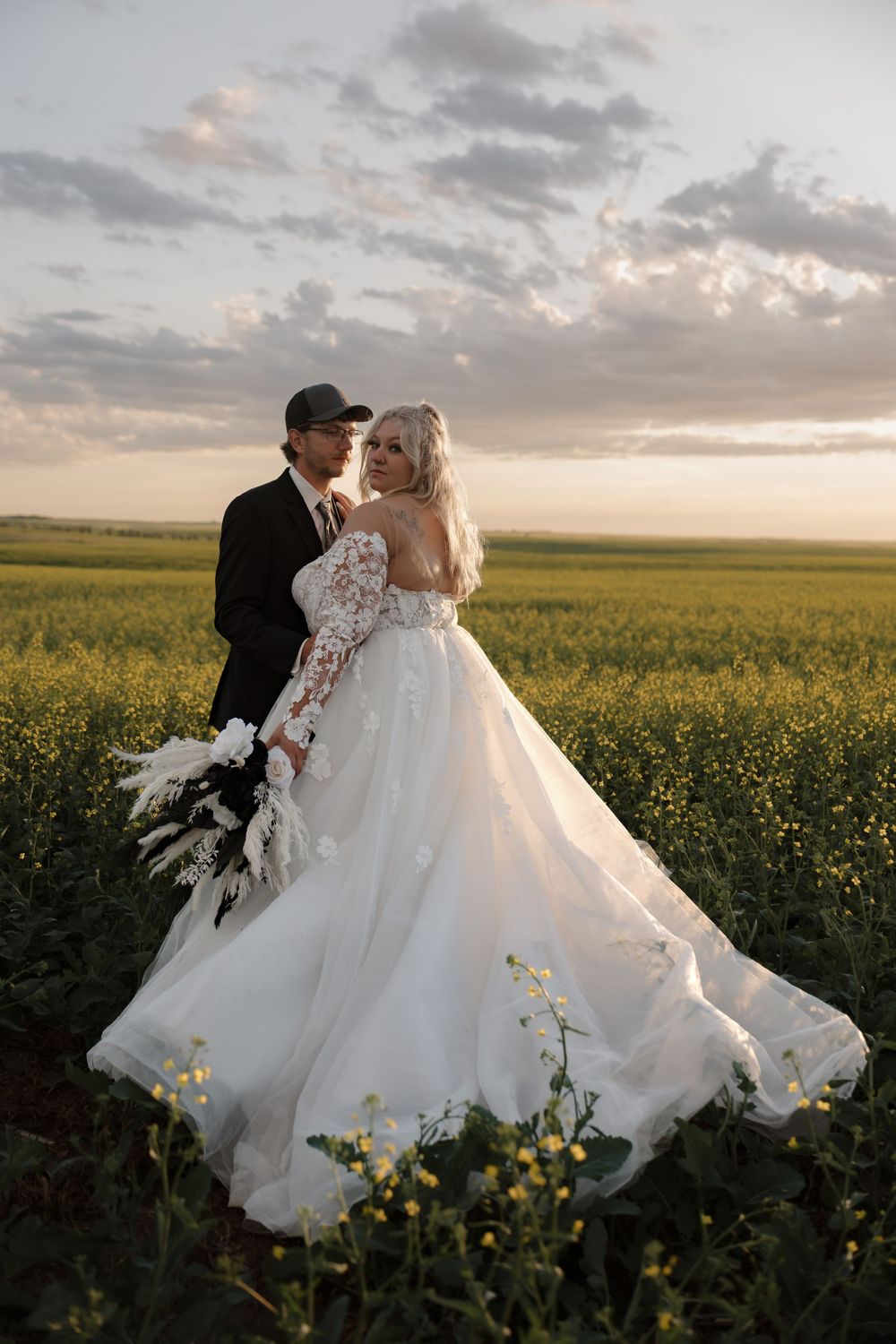 Bride in floral lace gown and groom in black suit embrace in golden canola field at sunset. Garrison, ND Photographer