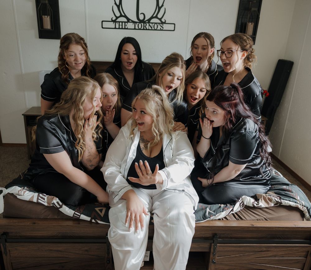 Bride in white pajamas surrounded by bridesmaids in black satin PJs on a bed during bridal party getting ready photos. Garrison, ND