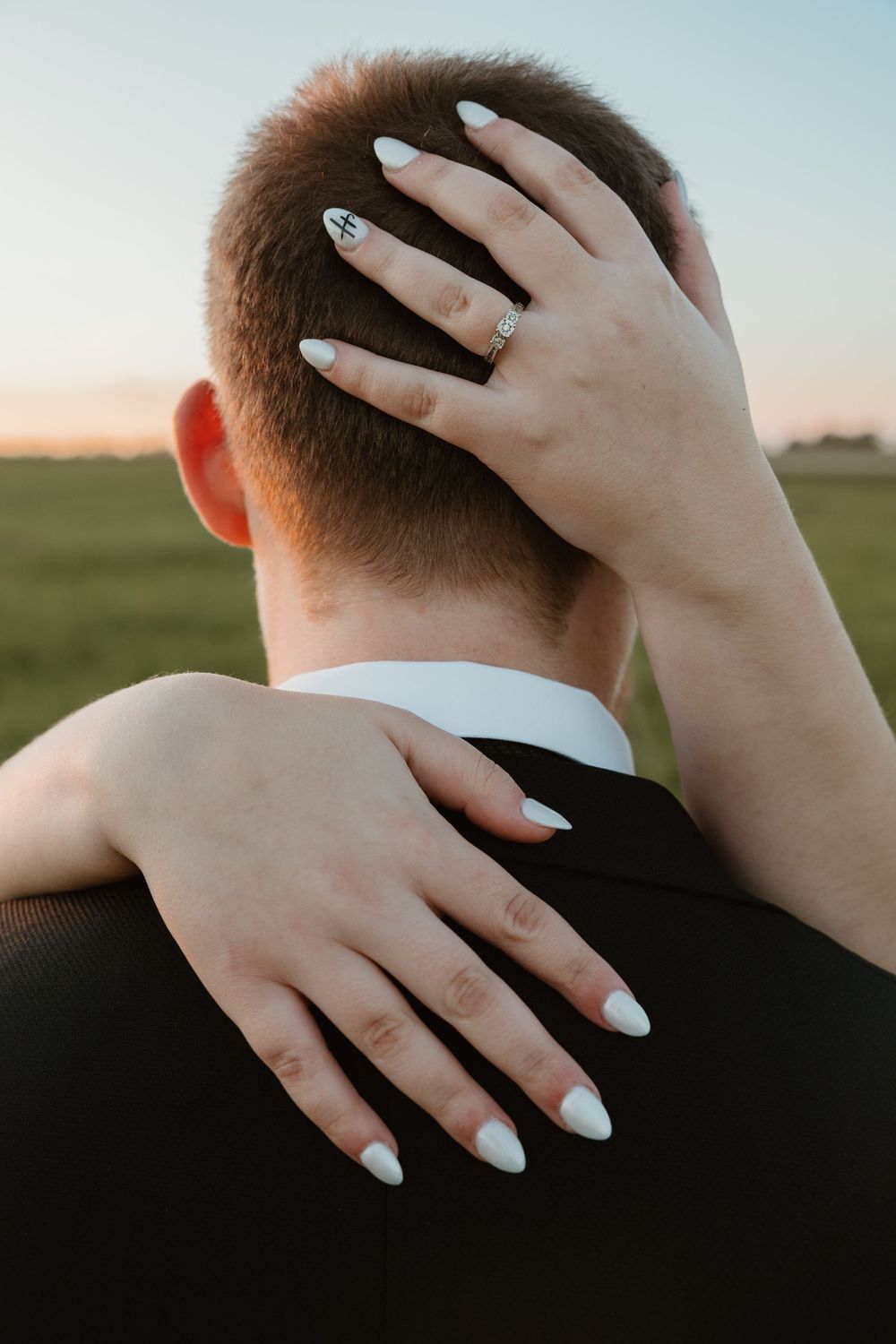 Woman with white manicure and engagement ring embracing a man in a suit from behind at sunset. Bismarck, ND travel photographer