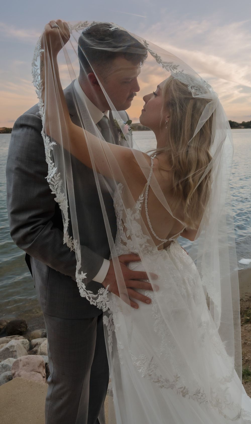 Bride and groom share romantic moment under lace veil by waterfront at sunset, bride in backless wedding gown. Bismarck, ND travel photo