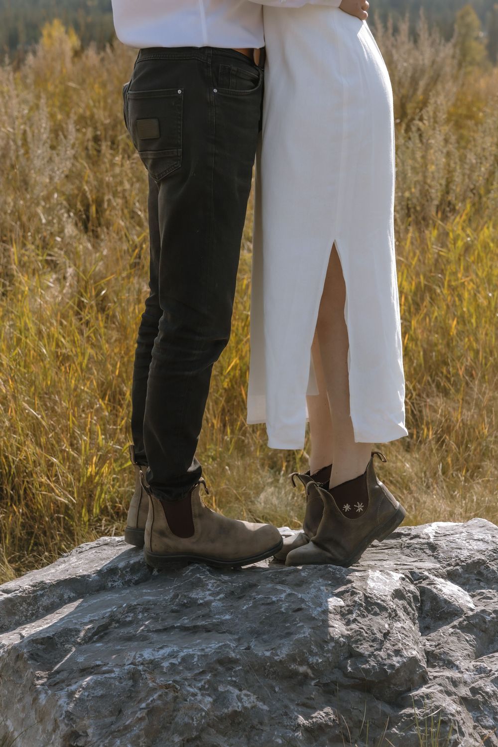 Couple standing on a rock in matching Chelsea boots, wearing white shirt and dress with black jeans in autumn field. Bismarck, ND travel