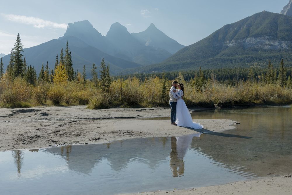 Bride and groom embrace on a sandy riverbank with stunning mountain peaks and autumn trees reflected in calm water. Bismarck, ND travel