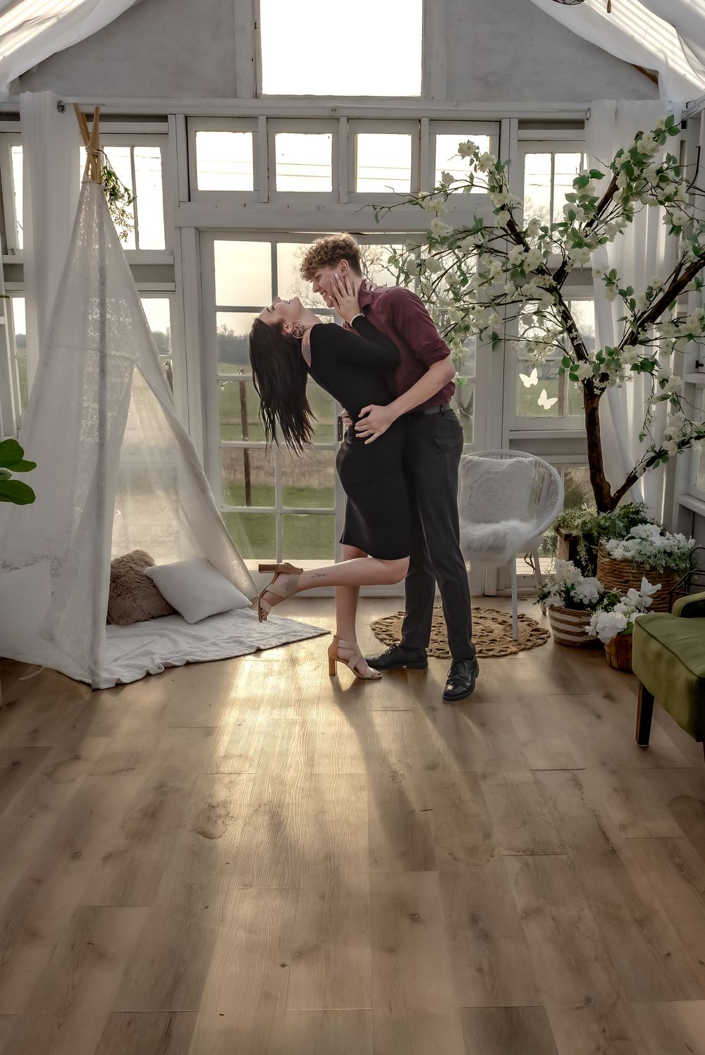 Couple sharing a romantic kiss in a sunlit greenhouse room with white teepee, blooming tree, and hardwood floors. Bismarck, ND photographer