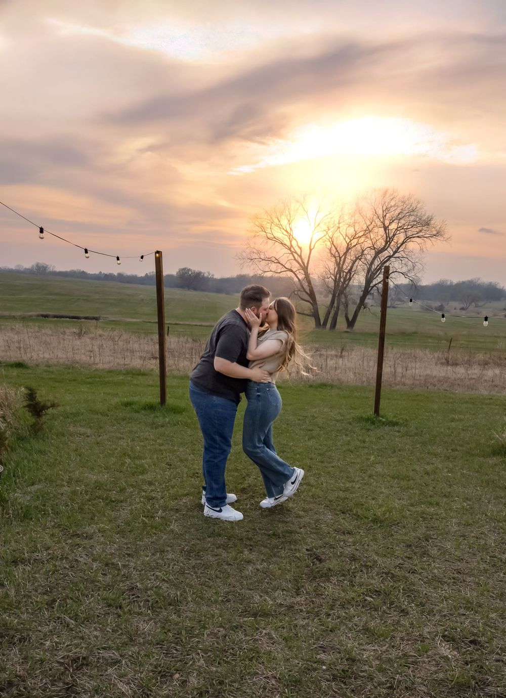 Couple sharing a romantic kiss at sunset in a rustic field with string lights and bare trees in the background. Bismarck, ND photographer