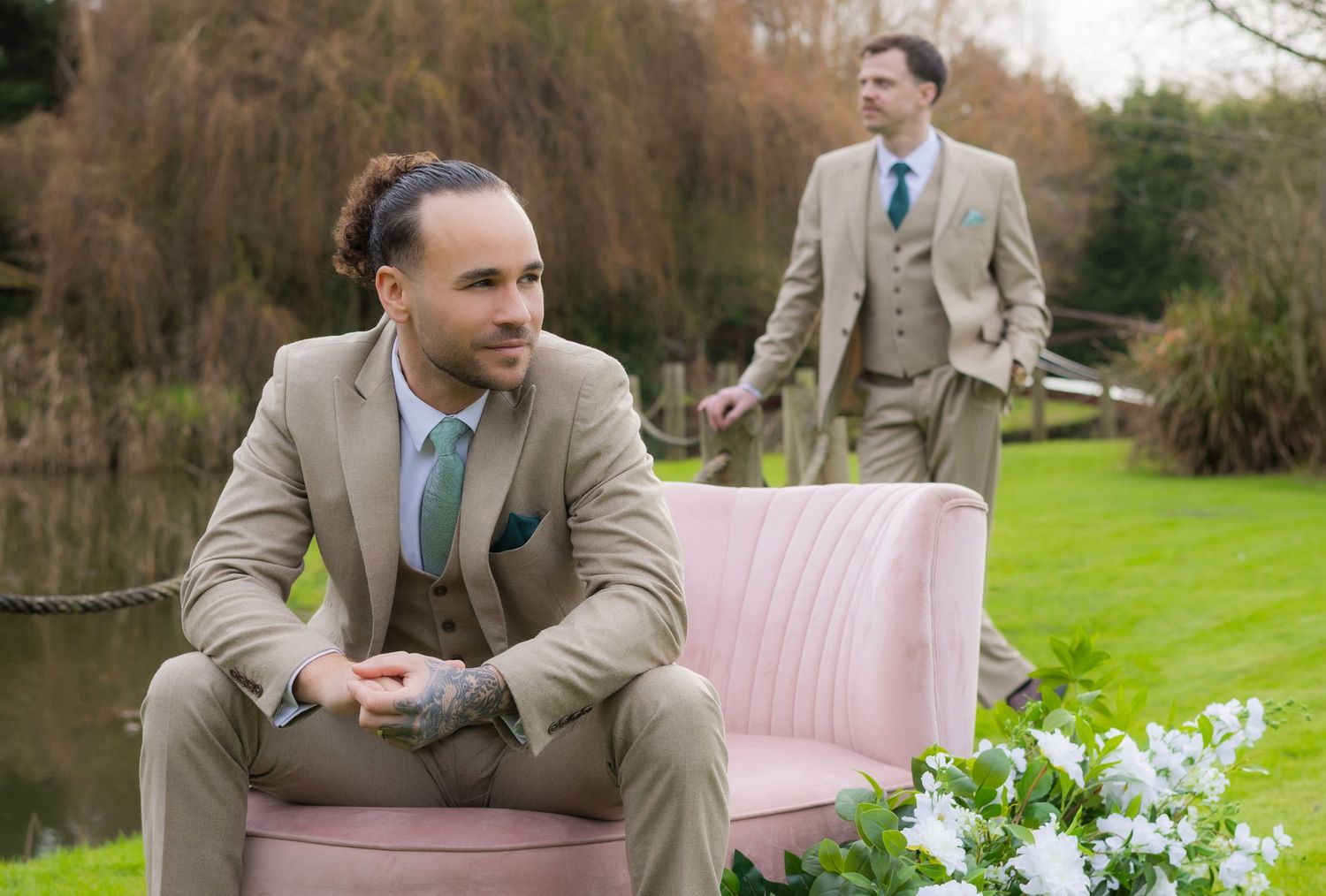 Two grooms in matching beige suits pose outdoors by a lake with a pink velvet sofa and white floral arrangement.