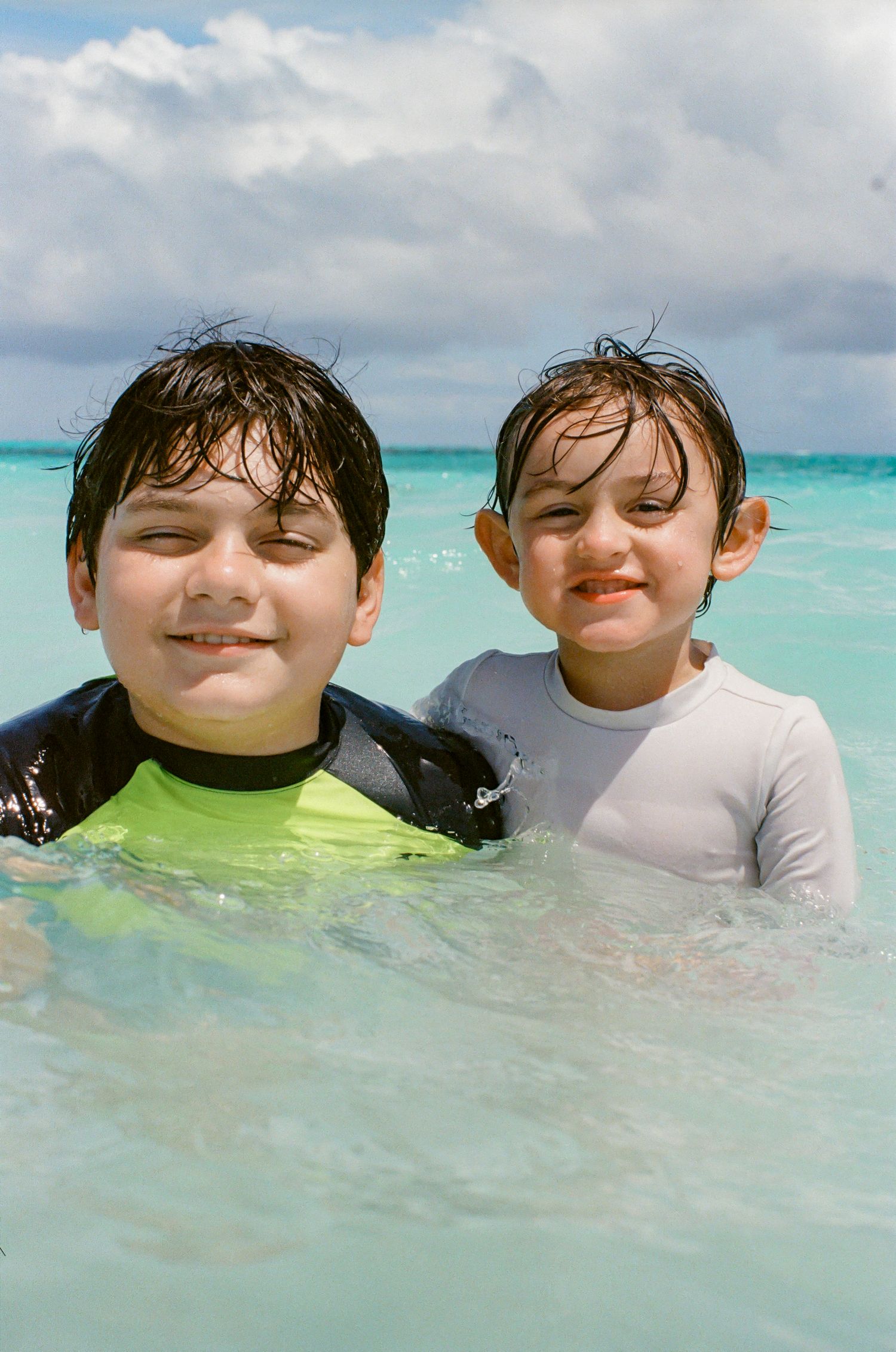 Two young boys smiling in turquoise ocean water, wearing rash guards, enjoying a sunny beach day.