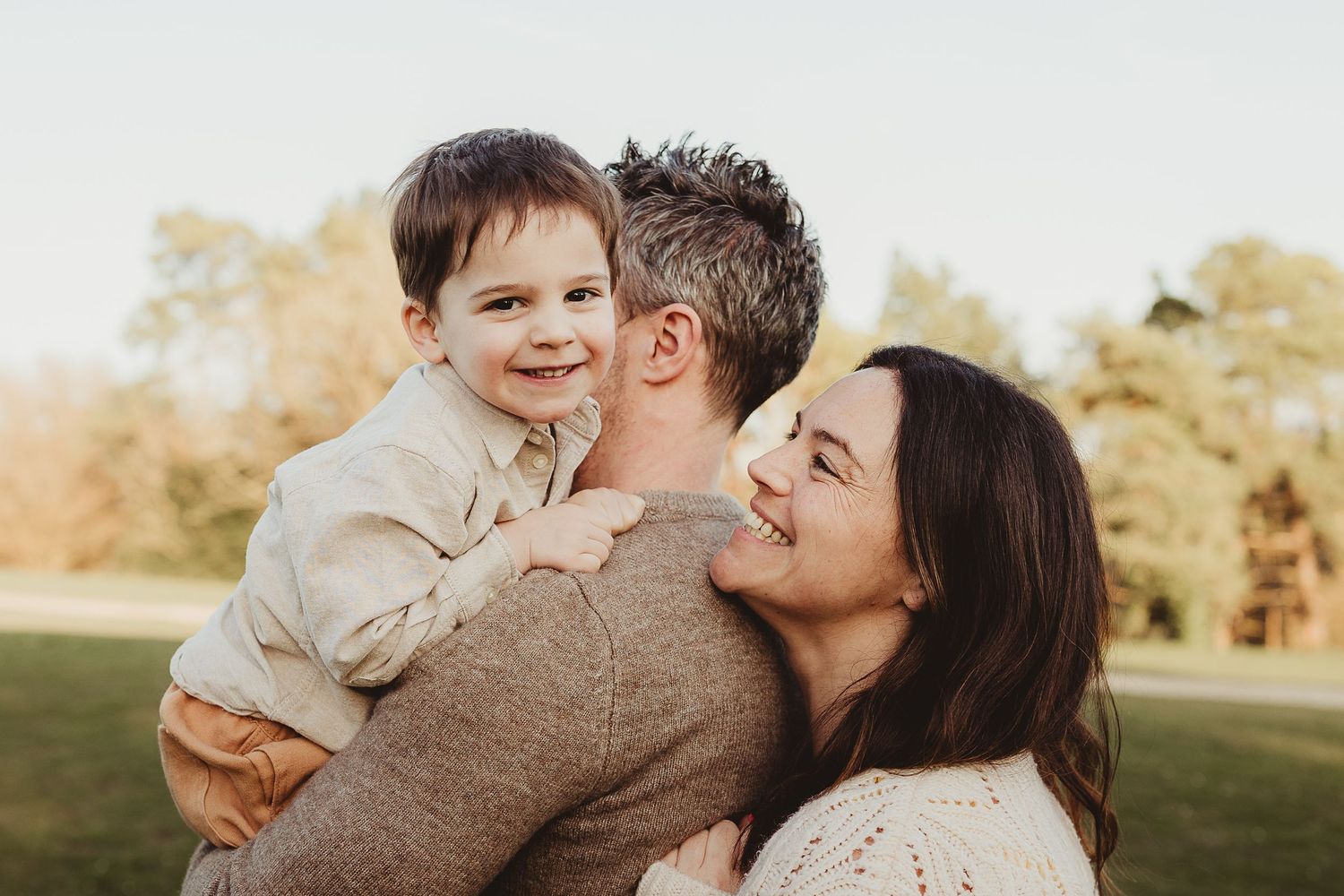 Happy family moment with parents kissing and hugging smiling toddler boy outdoors in a sunny park setting.