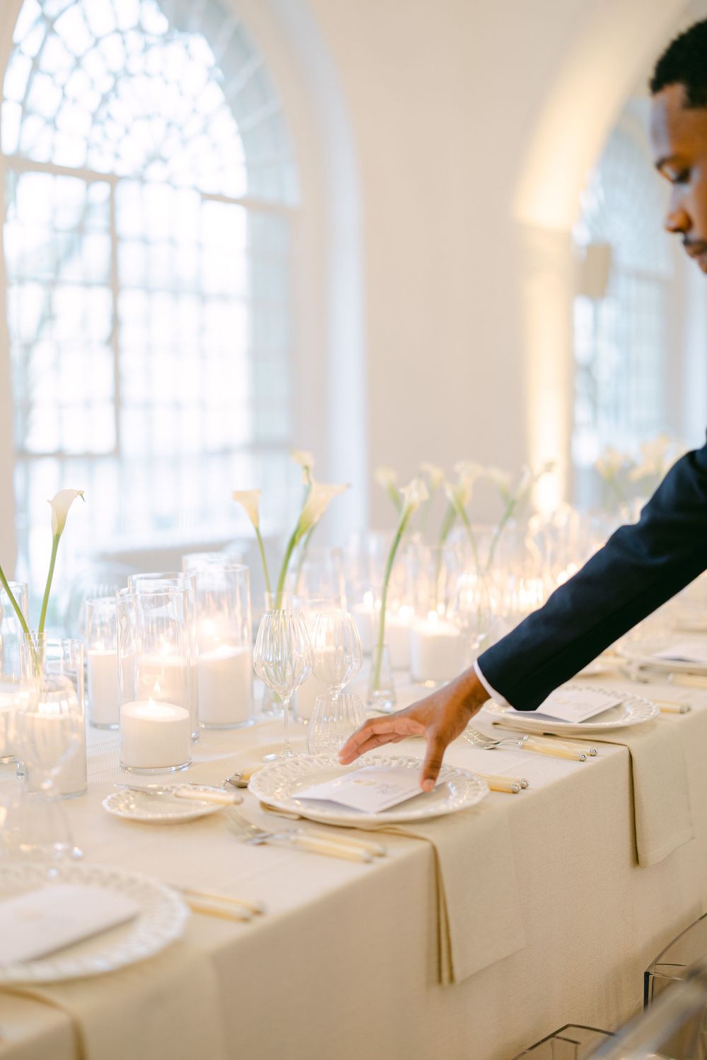 Server arranging white linen napkins on elegant wedding reception table with calla lilies and candles.