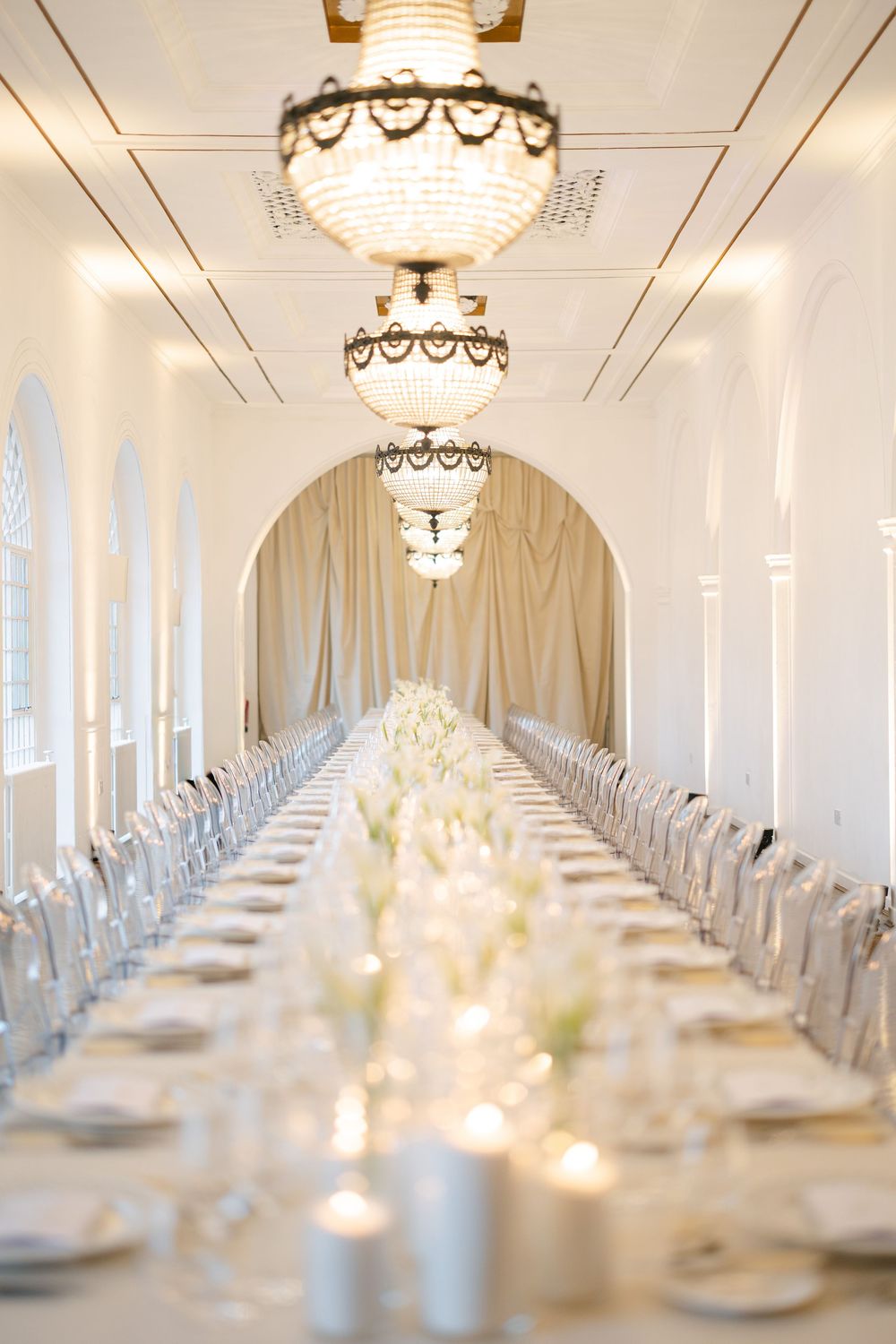 Elegant wedding reception hall with long white table, crystal chandeliers, candles, and white floral centerpieces.