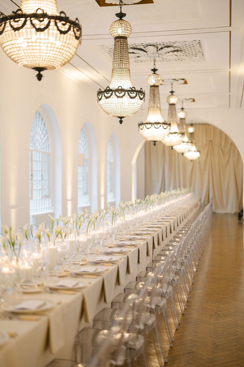 Elegant wedding reception hall with long dining tables, crystal chandeliers, white floral centerpieces, and ghost chairs in a grand venue.