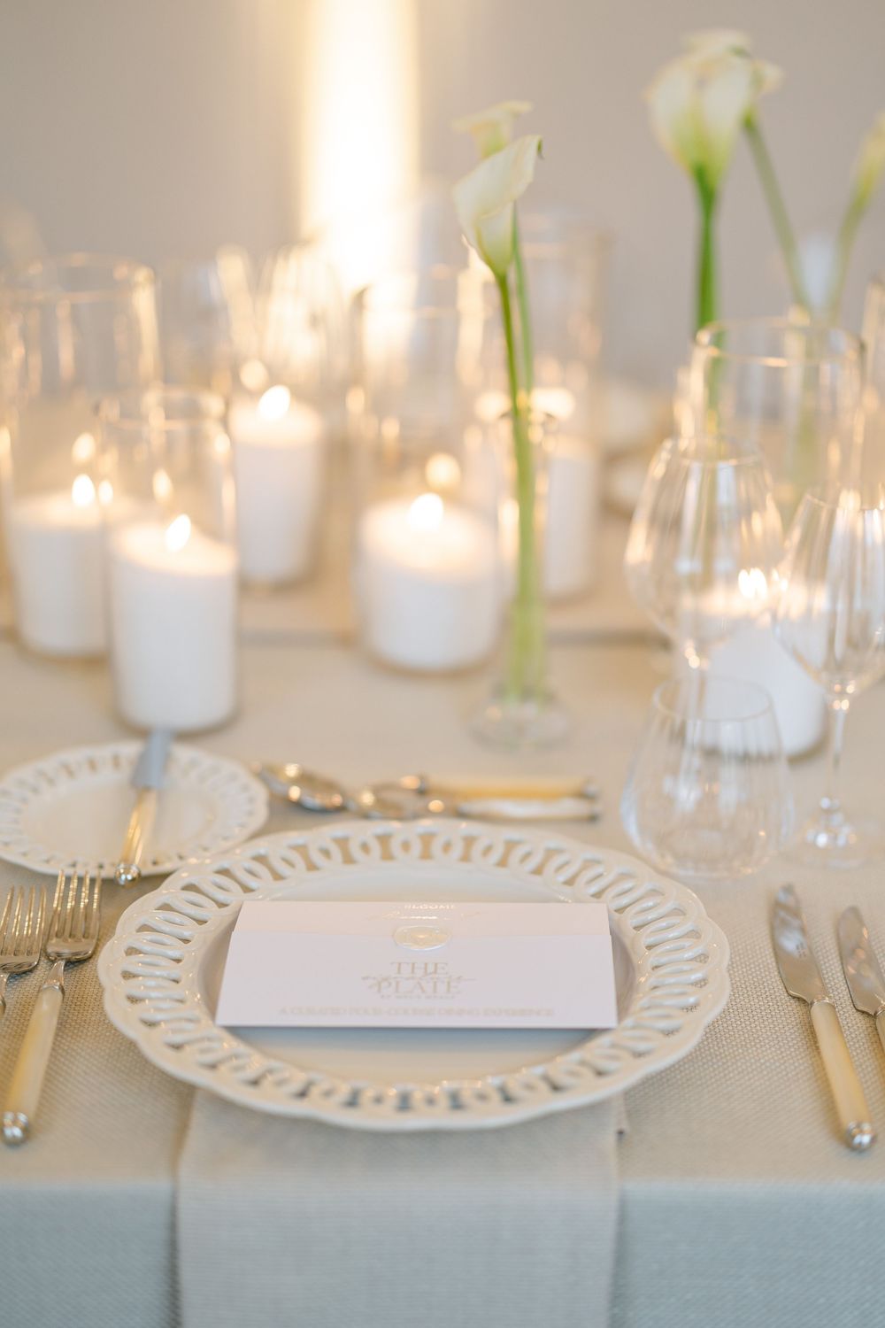 Romantic wedding reception place setting with white lattice charger plate, gold cutlery, and glowing candles on a neutral linen.
