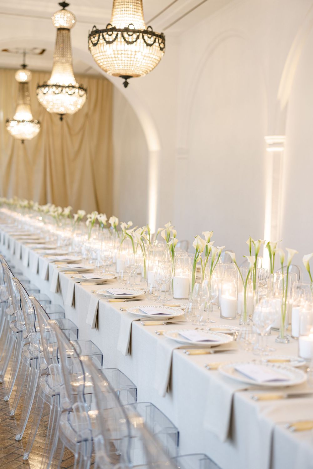 Elegant wedding reception with long white table, crystal chandeliers, ghost chairs, and white calla lily centerpieces.